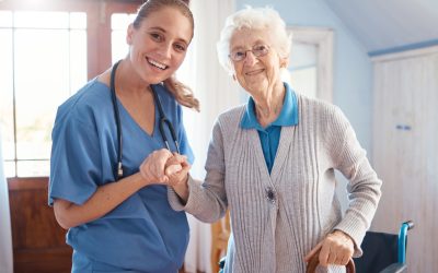 A Smiling Nurse In Blue Scrubs Holds Hands With An Elderly Assisted Living Resident