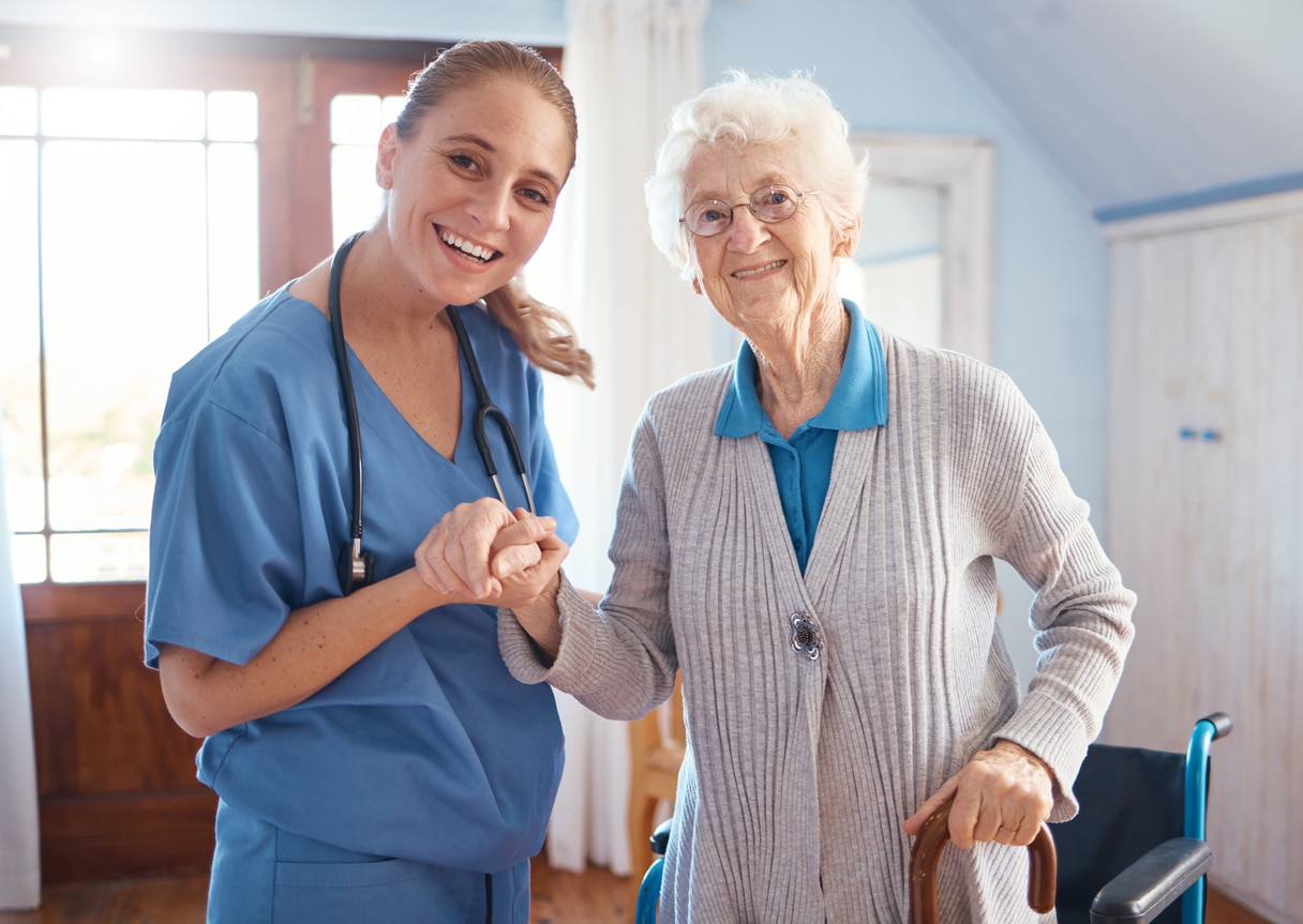 A smiling nurse in blue scrubs holds hands with an elderly assisted living resident