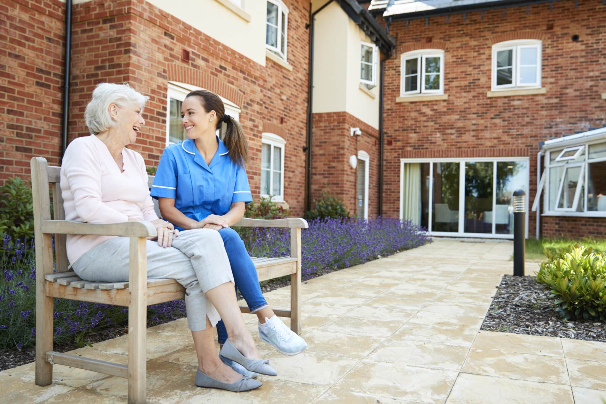 A senior and caretaker sitting on a bench just outside of a senior living facility