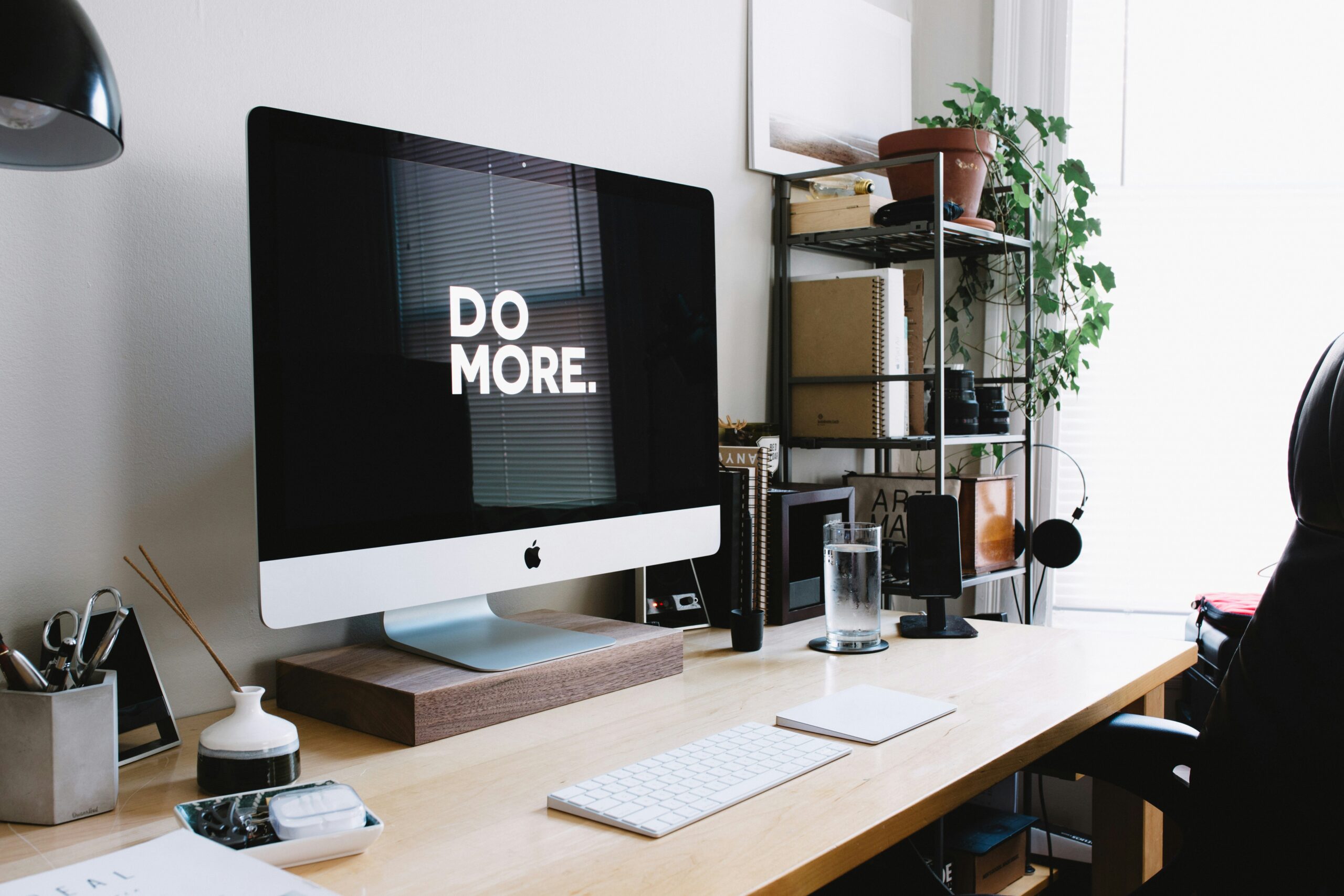 Desktop workspace featuring an Apple iMac displaying "DO MORE." text, with a keyboard, mouse, and various office supplies, emphasizing productivity in a digital marketing context.