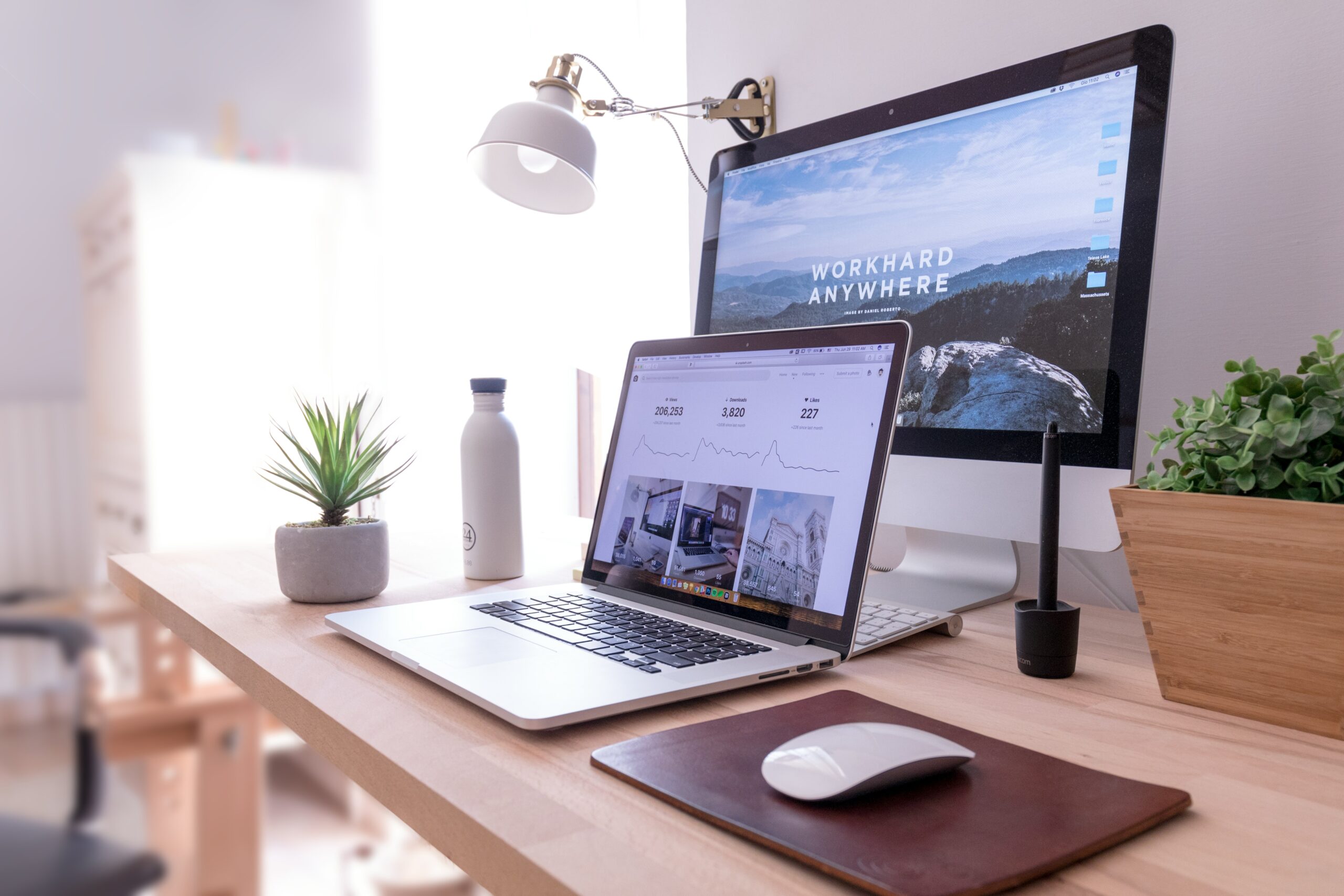 Desk setup with a laptop displaying analytics, a desktop monitor with "WORK HARD ANYWHERE" wallpaper, a small potted plant, a water bottle, and a mouse, emphasizing a modern workspace for digital marketing and HIPAA compliance discussions.